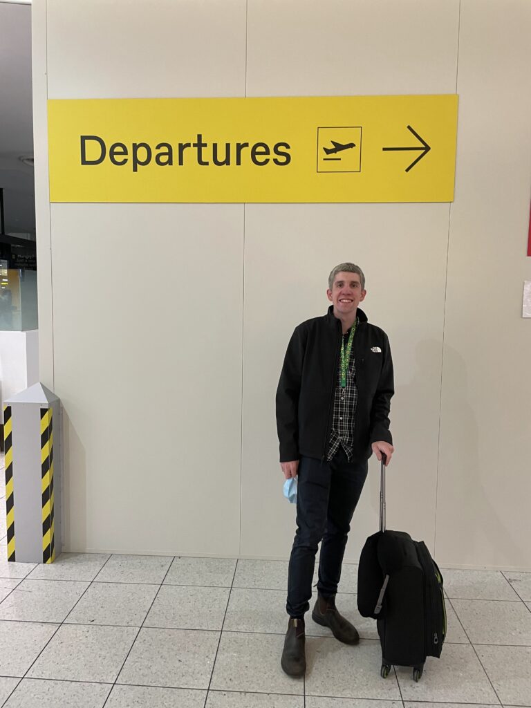 Jeremy standing in front of departures sign at Melbourne Airport with his suitcase. He is wearing his Northface Jacket a black cheque shirt and blue pants. 