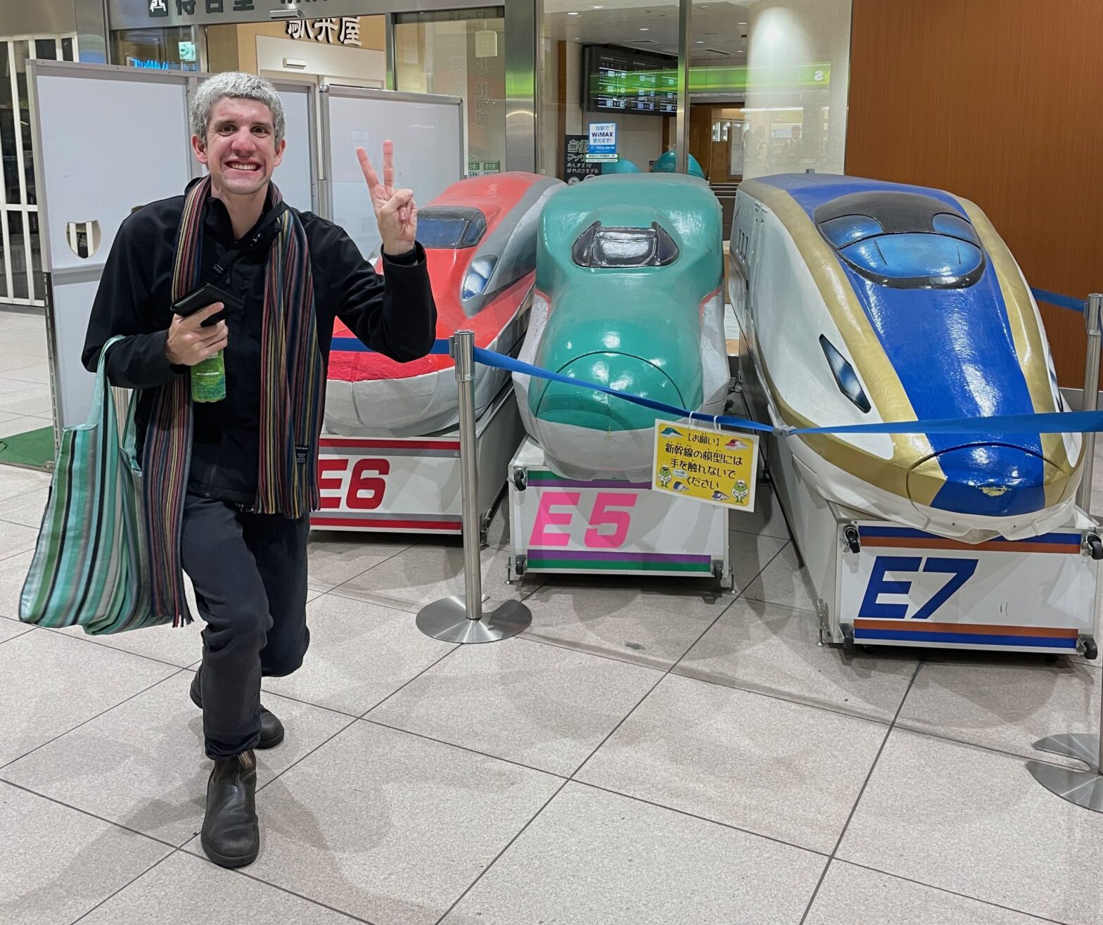 Jeremy wearing a black jacket and black pants with a rainbow scarf posing in front of models models of the different Shinkansen that go through Omiya. Pink E6, Green E5 and Blue E7.