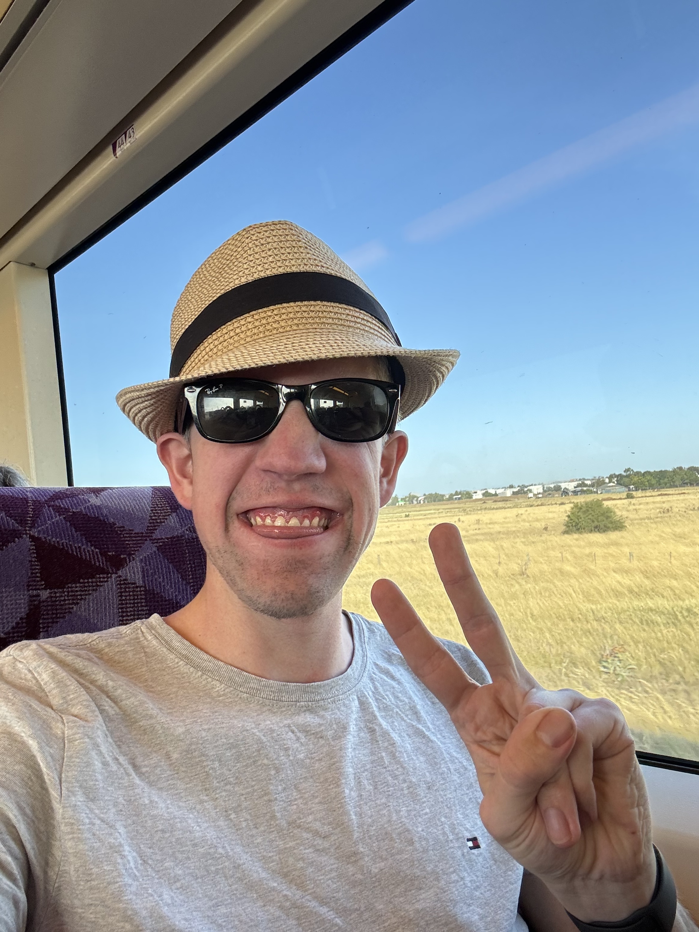 Jeremy wearing a grey t-shirt ad cream coloured hat with black band and sunglasses doing a peace sign. 