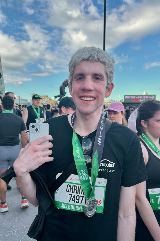 Jeremy smiling proudly whilst wearing the medal for completion of the 10km run. The sky is blue at the outside Gate 1 of the MCG